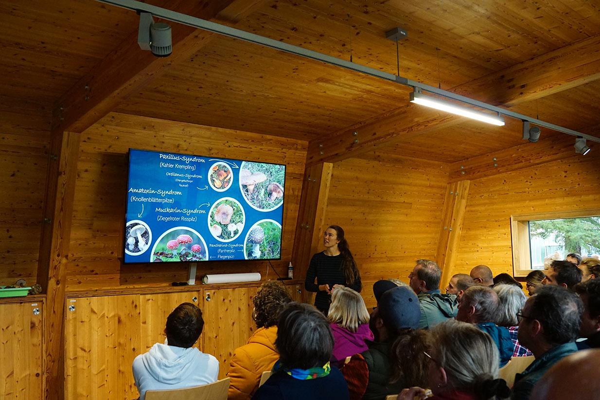 Foto vom Vortrag im holzverkleideten Zedernhaus, Referentin und Bildschirmfolie, davor Publikum